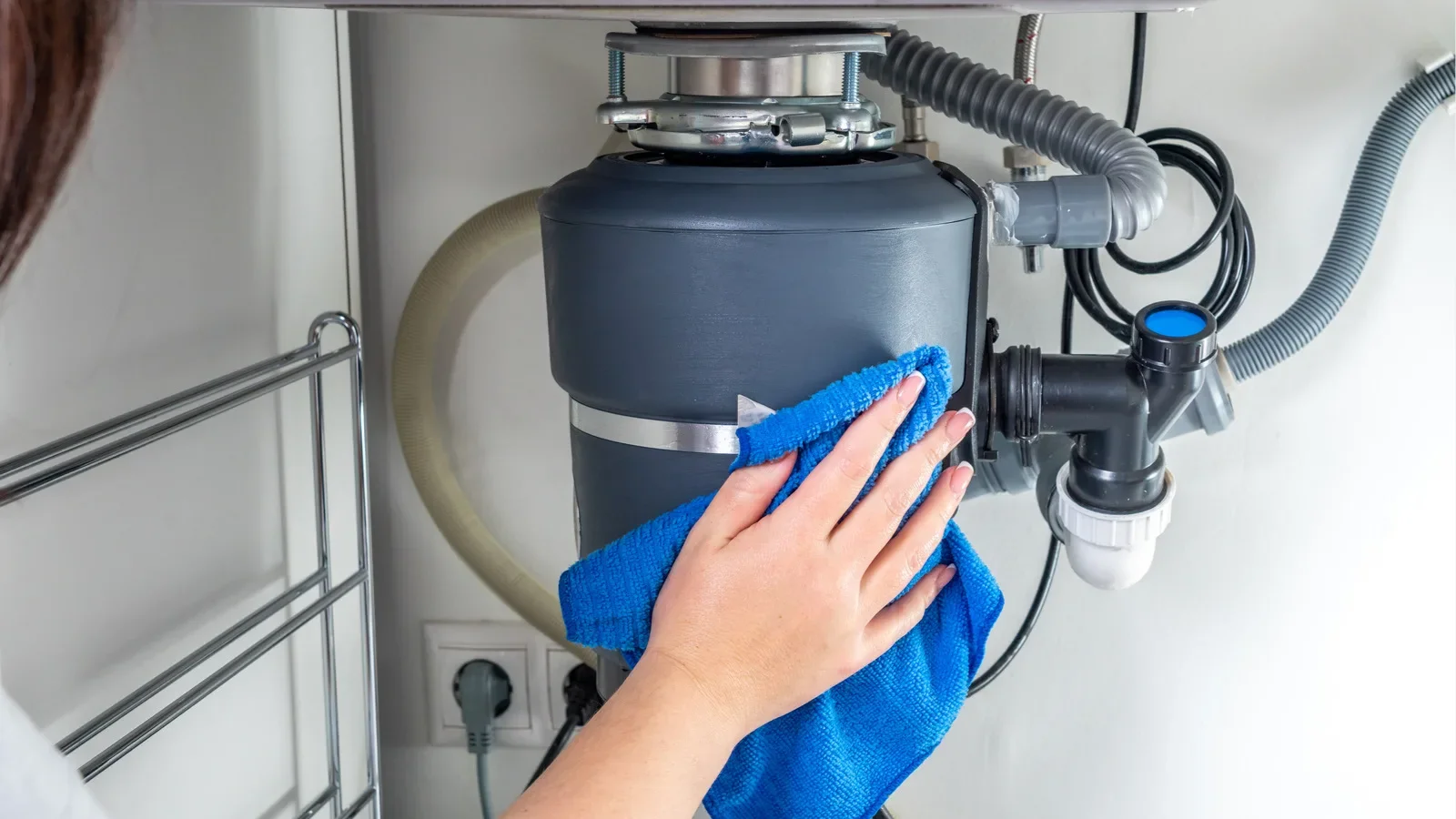 Hand cleaning garbage disposal unit under kitchen sink with blue cloth, showing plumbing connections and mounted motor assembly.