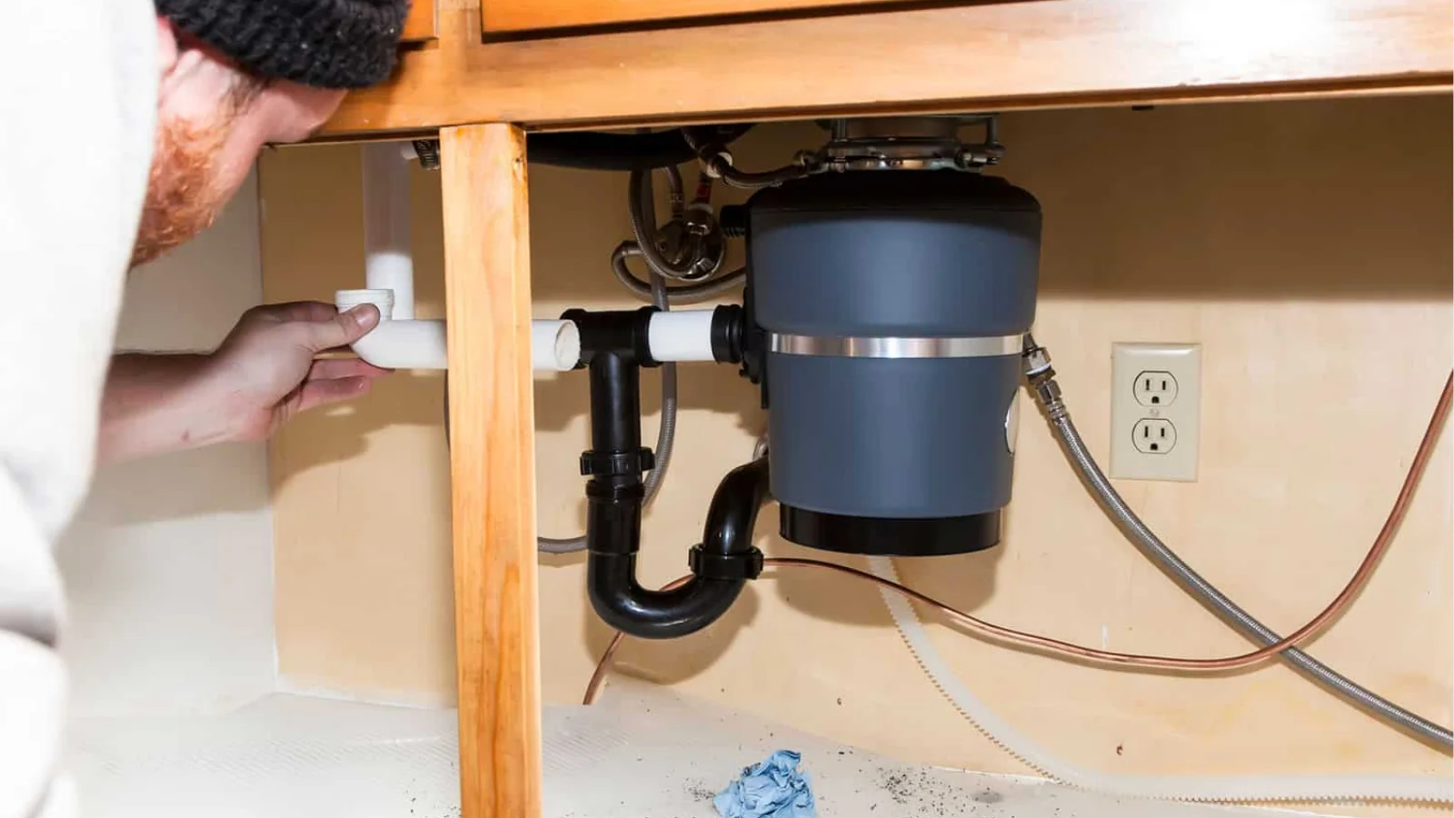 Person installing or adjusting PVC drain pipe connected to garbage disposal unit under kitchen sink cabinet with visible plumbing and outlet.